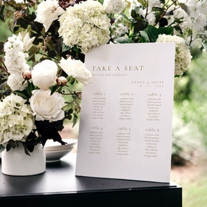 Puede incluir: Una tarjeta blanca con el texto "TAKE A SEAT" y "let's eat and celebrate!" en una fuente minimalista. Debajo del texto hay un plano de asientos para una boda con los nombres de los invitados enumerados debajo de cada número de mesa.