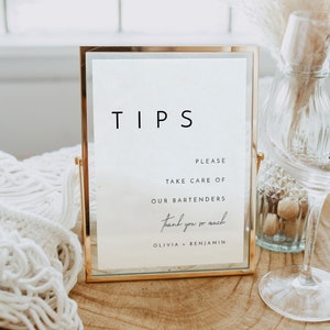 Könnte beinhalten: Ein goldfarbener Rahmen mit dem Wort "TIPS" in schwarzer Schrift. Das Schild enthält auch den Text "Bitte kümmern Sie sich um unsere Barkeeper. Vielen Dank. Olivia + Benjamin."
