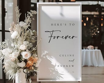 May include: White sign with a gold frame and stand, featuring the text "Here's to Forever" in black cursive script. Below the text, it reads "Celine and Andrew".