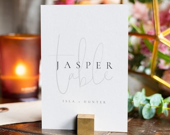 May include: White table number card with the word "Table" written in cursive script and the word "Jasper" in all caps. The card also includes the names "Isla" and "Hunter" in all caps.