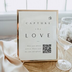 May include: A gold-framed sign with the text "CAPTURE the LOVE" and a QR code. The sign is on a wooden surface with a white fabric draped over it.