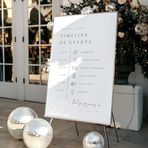 Peut inclure: Un panneau blanc pour un événement de mariage avec un cadre noir, affichant un calendrier des événements. Des boules disco argentées sont disposées autour du panneau. Le panneau indique "Claire + Lucas" et "Calendrier des événements".