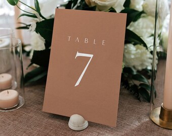 May include: A brown card with white text that reads "TABLE 7" on a table setting with white flowers and candles.