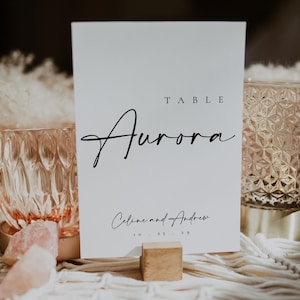 Könnte beinhalten: Weißes Tischschild mit dem Wort "Aurora" in eleganter Schrift. Das Schild enthält auch die Namen "Celine and Andrew" und das Datum "10.25.29". Das Schild steht auf einem Makramee-Tischläufer mit dekorativen Glas- und Rosenquarz-Akzenten.