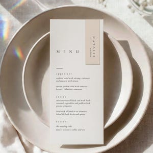 May include: A rectangular white paper menu card with black text featuring the names Natalie and Madden on a beige vertical accent strip. The menu lists appetizers, entrees, and desserts including seafood salad, black cod, and lamb.