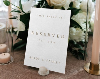 May include: A white card with gold lettering that reads "This table is reserved for the bride's family". The card is on a table with a white tablecloth and a floral centerpiece.