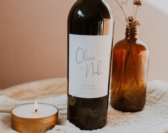 May include: A wine bottle with a white label that says "Olivia + Noah" and "08.09.2023 Let's celebrate!"  The bottle is on a wooden surface with a white cloth and a lit candle.