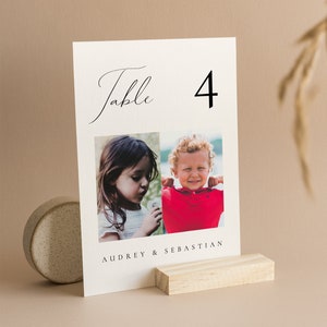 Può includere: Un biglietto numerico da tavolo color crema con la parola "Table" in corsivo e il numero "4" in grassetto. Il biglietto presenta due foto di bambini e i nomi "Audrey & Sebastian". Il biglietto è tenuto da un piccolo blocco di legno.
