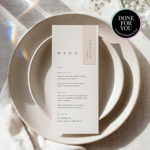 May include: A vertical rectangular menu card printed on off-white paper with black text, featuring the word MENU, the name Natalie Madden, and a list of appetizers, entrees, and desserts. The card is centered on a stack of two round ceramic plates.