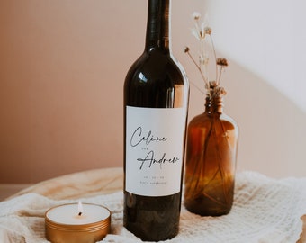 May include: A wine bottle with a white label that says "Celine and Andrew" with a wedding date and the words "Let's celebrate!" A small candle and a brown glass bottle with dried flowers are on a wooden tray with a white cloth.