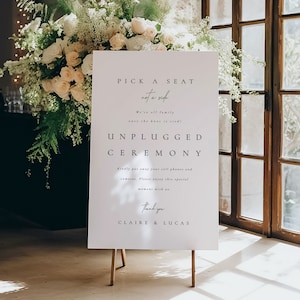 May include: A white sign on a wooden easel with the text "PICK A SEAT not a side. UNPLUGGED CEREMONY." The sign is surrounded by floral arrangements. The sign requests guests to put away cell phones and cameras.