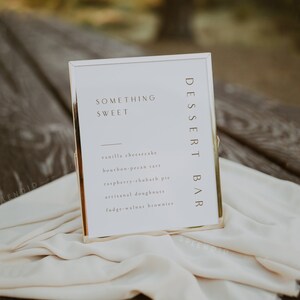 May include: A gold-framed sign with the text "Something Sweet" and a list of desserts: vanilla cheesecake, bourbon-pecan tart, raspberry-rhubarb pie, artisanal doughnuts, and fudge-walnut brownies. The sign is on a white fabric surface.