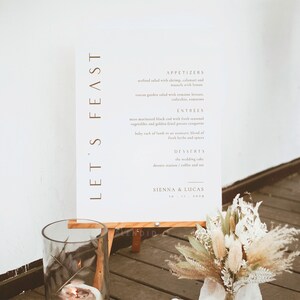 Könnte beinhalten: Ein weißes Schild mit braunem Text auf einem Holzständer. Das Schild lautet "Let's Feast" und listet ein Hochzeitsmenü mit Vorspeisen, Hauptgerichten, Desserts und den Namen Sienna & Lucas mit dem Datum 10.21.2029.