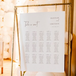 May include: A wedding seating chart with the words "Take a seat" in elegant script. The chart lists table numbers and guest names, displayed on a white background within a gold metal frame. The names "Hannah and Greyson" and the date "10.05.21" are also displayed.