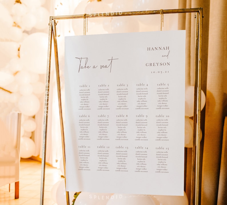 May include: A wedding seating chart with the words "Take a seat" in elegant script. The chart lists table numbers and guest names, displayed on a white background within a gold metal frame. The names "Hannah and Greyson" and the date "10.05.21" are also displayed.