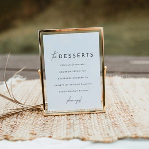 May include: A gold-framed sign with a white background and black text that reads "The Desserts" and lists a variety of desserts, including vanilla cheesecake, bourbon pecan tart, raspberry rhubarb pie, artisan doughnuts, and fudge walnut brownies. The sign is on a woven mat.