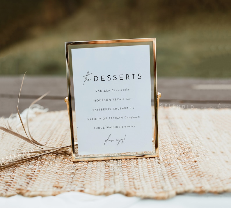 May include: A gold-framed sign with a white background and black text that reads "The Desserts" and lists a variety of desserts, including vanilla cheesecake, bourbon pecan tart, raspberry rhubarb pie, artisan doughnuts, and fudge walnut brownies. The sign is on a woven mat.