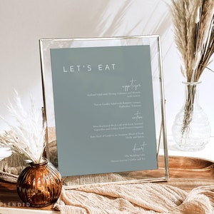 May include: A framed menu with a blue background and white text. The menu is titled "Let's Eat" and lists appetizers, entrees, and desserts. The menu is displayed on a wooden table with a vase of dried flowers.