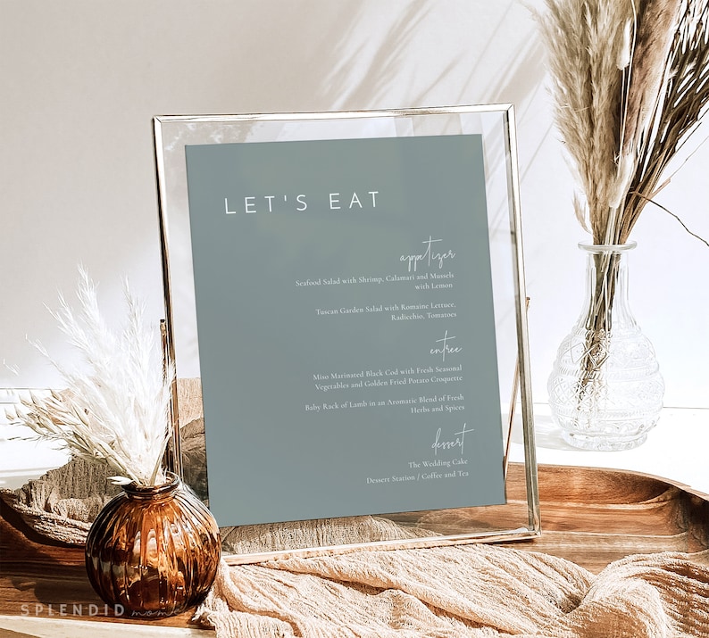 May include: A framed menu with a blue background and white text. The menu is titled "Let's Eat" and lists appetizers, entrees, and desserts. The menu is displayed on a wooden table with a vase of dried flowers.