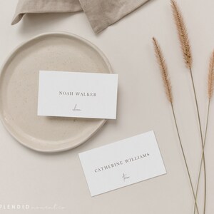 May include: Two white place cards with names printed in a minimalist font. One card reads "Noah Walker" with "forever" written below. The other card reads "Catherine Williams" with "forever" below. A beige plate, linen, and dried grass complete the elegant arrangement.