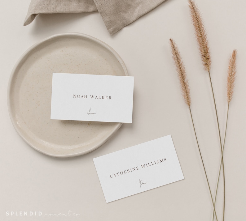 May include: Two white place cards with names printed in a minimalist font. One card reads "Noah Walker" with "forever" written below. The other card reads "Catherine Williams" with "forever" below. A beige plate, linen, and dried grass complete the elegant arrangement.