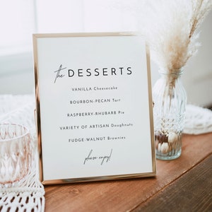 May include: A gold-framed sign with the word "DESSERTS" in cursive script. The sign lists a variety of desserts, including vanilla cheesecake, bourbon-pecan tart, raspberry-rhubarb pie, artisan doughnuts, and fudge-walnut brownies. The sign ends with the phrase "please enjoy!"
