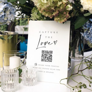 May include: White sign with black text that reads "Capture the Love" with a heart symbol. A black and white QR code is below the text. The text below the QR code reads "Share your photos with us! Please scan the code & upload."