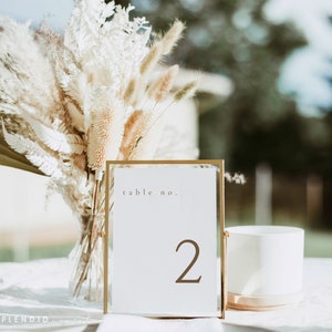 May include: A gold-framed table number card with the number "2" in brown. The card is on a white surface, with a vase of dried flowers and a white cylindrical object in the background. The text "table no." is at the top.