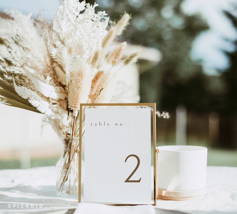 May include: A gold-framed table number card with the number "2" in brown. The card is on a white surface, with a vase of dried flowers and a white cylindrical object in the background. The text "table no." is at the top.
