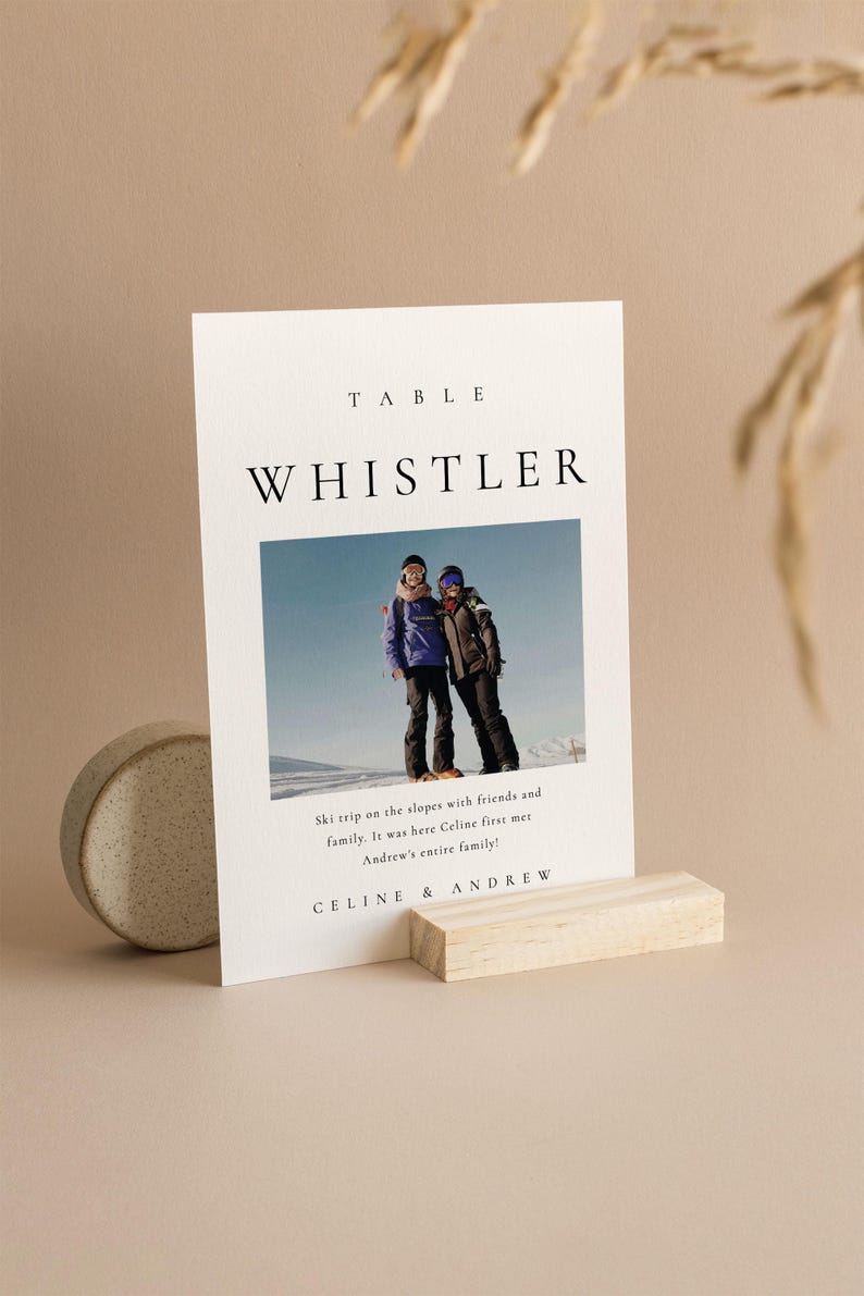 May include: A white table card with black text that reads "TABLE WHISTLER". A photo of two people in winter clothing on a ski slope is below the text. The text below the photo reads "Ski trip on the slopes with friends and family. It was here Celine first met Andrew's entire family!" and "CELINE & ANDREW".