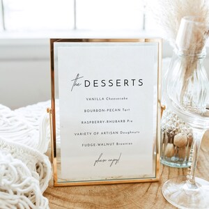 May include: A gold-framed sign with a white background lists a dessert menu: Vanilla Cheesecake, Bourbon-Pecan Tart, Raspberry-Rhubarb Pie, Variety of Artisan Doughnuts, Fudge-Walnut Brownies. The sign reads "the DESSERTS" and "please enjoy!"