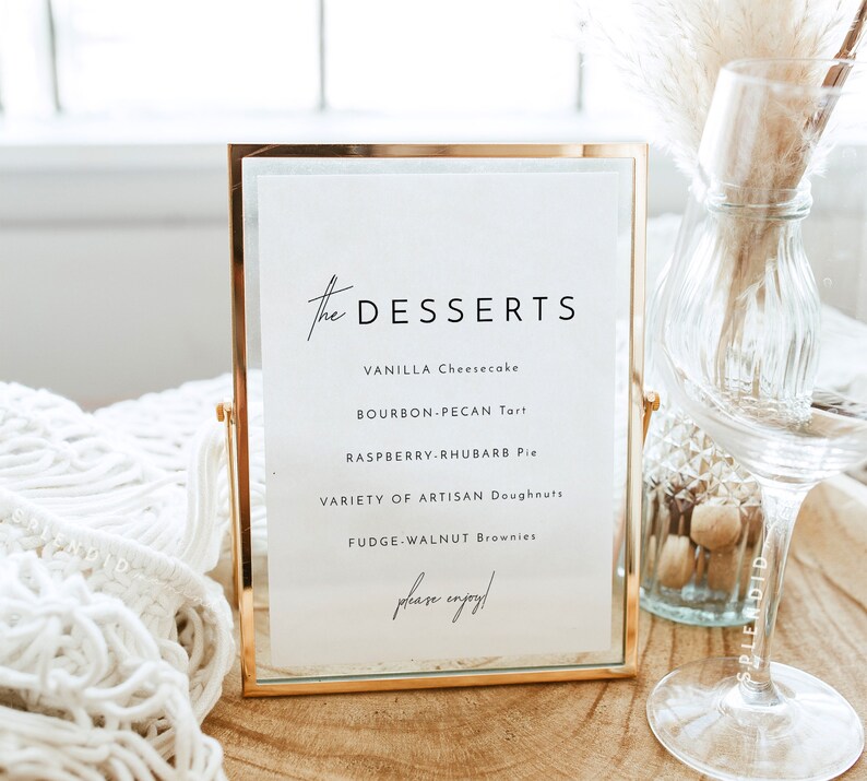 May include: A gold-framed sign with a white background lists a dessert menu: Vanilla Cheesecake, Bourbon-Pecan Tart, Raspberry-Rhubarb Pie, Variety of Artisan Doughnuts, Fudge-Walnut Brownies. The sign reads "the DESSERTS" and "please enjoy!"
