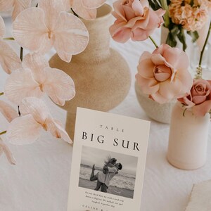 May include: A white table number card with the text "TABLE BIG SUR" in black font. The card features a black and white photo of a couple on a beach. The text below the photo reads "On our third date we took a road trip down to Big Sur. We walked, ate and laughed. A perfect day. CELINE & ANDREW". The card is sitting on a gold metal stand.