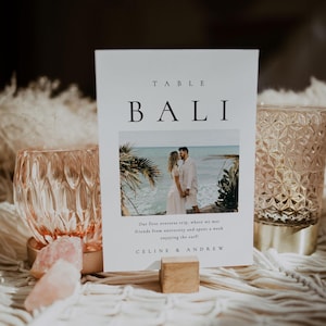 May include: A white table number card with the text "TABLE BALI" in black font. The card features a photo of a couple standing on a beach in Bali, with the text "Our first overseas trip, where we met friends from university and spent a week enjoying the surf!" and "CELINE & ANDREW" below the photo.