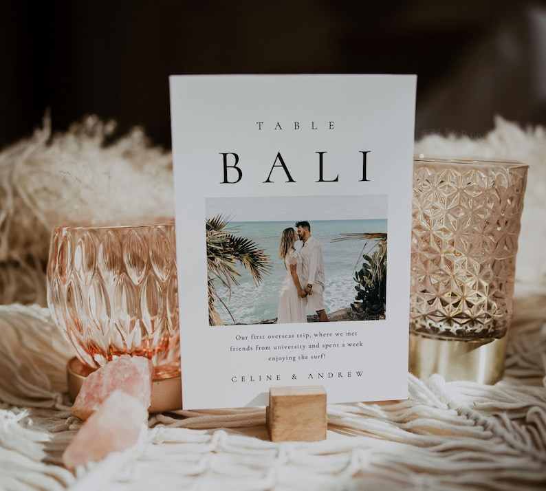 May include: A white table number card with the text "TABLE BALI" in black font. The card features a photo of a couple standing on a beach in Bali, with the text "Our first overseas trip, where we met friends from university and spent a week enjoying the surf!" and "CELINE & ANDREW" below the photo.