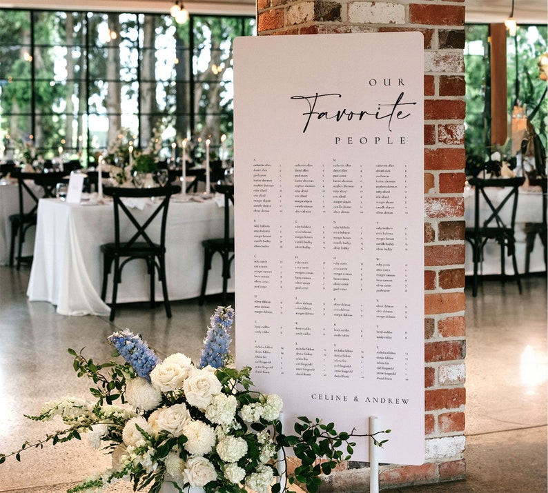 May include: A white wedding seating chart with the text "Our Favorite People" and "Celine & Andrew" at the bottom. The chart is displayed on a stand in front of a brick wall.