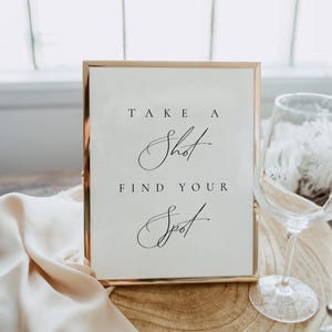 May include: A gold-framed sign with the text "Take a Shot Find Your Spot" in black calligraphy. The sign is sitting on a wooden surface with a white cloth and a clear wine glass.