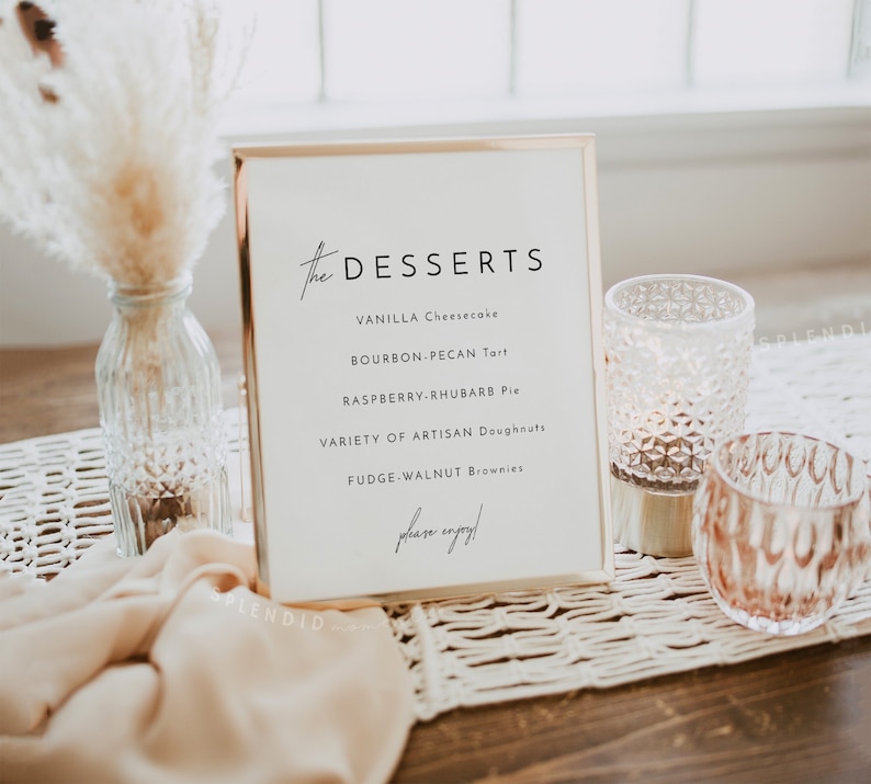 May include: A gold framed sign with the text "the DESSERTS" and a list of desserts: Vanilla Cheesecake, Bourbon-Pecan Tart, Raspberry-Rhubarb Pie, Variety of Artisan Doughnuts, Fudge-Walnut Brownies. The sign is on a table with a woven placemat and two glass candle holders.
