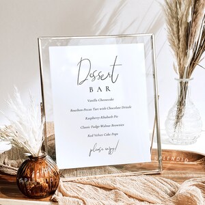 Puede incluir: Un cartel con marco dorado, fondo blanco y texto negro que dice "Dessert Bar". El cartel enumera una variedad de postres, incluyendo pastel de queso de vainilla, tarta de bourbon y nuez pecana con glaseado de chocolate, pastel de frambuesa y ruibarbo, brownies cl&aacute;sicos de fudge y nuez, y cake pops de terciopelo rojo. El cartel tambi&eacute;n incluye el texto "please enjoy!"