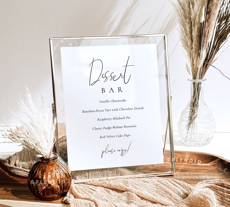 Puede incluir: Un cartel con marco dorado, fondo blanco y texto negro que dice "Dessert Bar". El cartel enumera una variedad de postres, incluyendo pastel de queso de vainilla, tarta de bourbon y nuez pecana con glaseado de chocolate, pastel de frambuesa y ruibarbo, brownies cl&aacute;sicos de fudge y nuez, y cake pops de terciopelo rojo. El cartel tambi&eacute;n incluye el texto "please enjoy!"