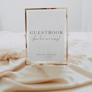 May include: A gold-framed sign with the words "GUESTBOOK" and "please leave us a message!" in elegant script. Below, the names "HANNAH + GREYSON" and the date "10/27/2022" are displayed. The sign is set on a soft, cream-colored surface.