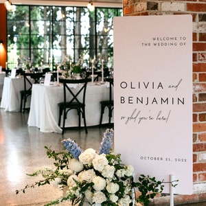 Può includere: Un cartello di matrimonio bianco con testo nero che recita "Benvenuti al matrimonio di Olivia e Benjamin, siamo felici che siate qui!" e "25 ottobre 2022".