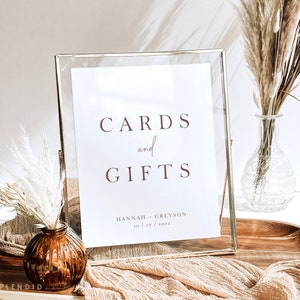 May include: A clear glass frame holds a white sign with the words "CARDS and GIFTS" in brown lettering. The sign also includes names and a date. Decorative vases with dried flowers are in the background, creating a rustic aesthetic.