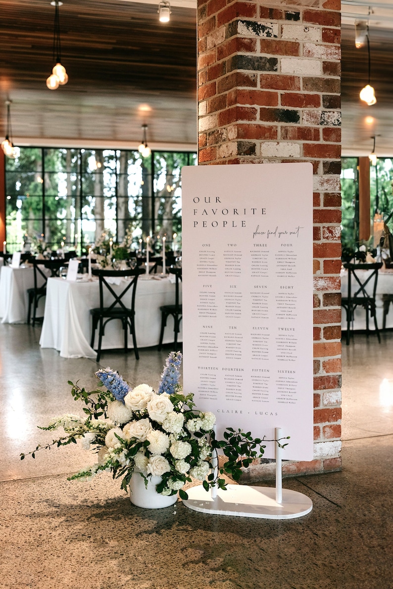 May include: A white sign with the text "Our Favorite People" and a seating chart for a wedding reception. The sign is on a white stand and is in front of a brick wall.