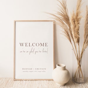 May include: A framed welcome sign with the text "WELCOME we're so glad you're here!" in brown lettering. The sign includes the names "HANNAH + GREYSON" and the date "saturday, august 11th / napa valley". The sign is next to a vase with dried plants.