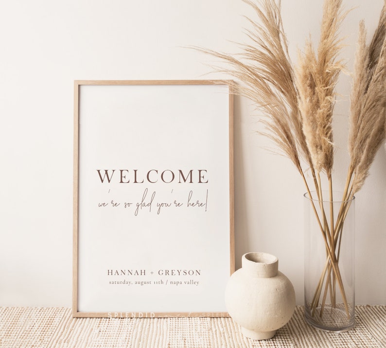 May include: A framed welcome sign with the text "WELCOME we're so glad you're here!" in brown lettering. The sign includes the names "HANNAH + GREYSON" and the date "saturday, august 11th / napa valley". The sign is next to a vase with dried plants.