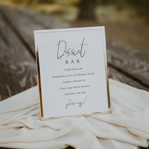 Puede incluir: Un cartel con marco dorado, fondo blanco y texto negro que dice "Dessert Bar". El cartel enumera una variedad de postres, que incluyen pastel de queso de vainilla, tarta de bourbon y nuez pecana con llovizna de chocolate, pastel de frambuesa y ruibarbo, brownies cl&aacute;sicos de fudge y nuez y cake pops de terciopelo rojo. El cartel tambi&eacute;n incluye la frase "&iexcl;por favor, disfruta!"