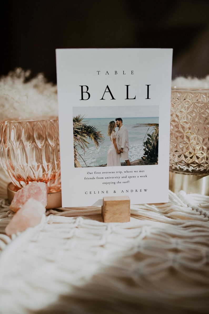 May include: A white card with black text that reads "TABLE BALI" and a photo of a couple on a beach. The text below the photo reads "Our first overseas trip, where we met friends from university and spent a week enjoying the surf! CELINE & ANDREW"