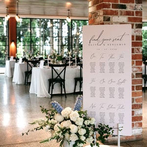 May include: A white sign with black text that reads "find your seat Olivia & Benjamin 10.12.2029" with a list of names under each table number. The sign is standing on a white stand in front of a brick wall. A white vase with a bouquet of white and blue flowers is in the foreground.