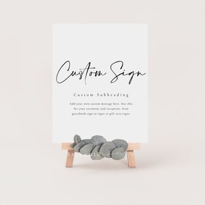 May include: A white sign with black text that reads "Custom Sign" and "Custom Subheading". The text below the subheading reads "Add your own custom message here. Use this for your ceremony and reception, from guestbook sign-in signs to gift area signs."